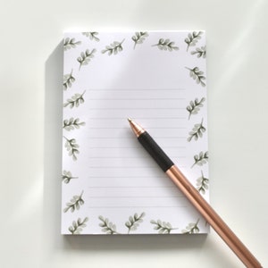 Op de afbeelding: Een wit notitieboekje met een groene bladrand en gestreept papier. Een roosgoudkleurige pen ligt op het notitieboekje.