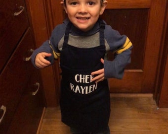 Könnte beinhalten: Ein Kind trägt eine weiße Kochmütze und eine marineblaue Schürze, beide mit dem Namen "CHEF RAYLEN" in Weiß aufgedruckt. Das Kind trägt außerdem einen blau-gelben Pullover, Jeans und gemusterte Schuhe. Das Kind lächelt.
