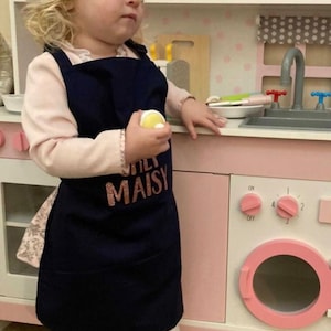 K&ouml;nnte beinhalten: Ein kleines Kind tr&auml;gt eine wei&szlig;e Kochm&uuml;tze und eine marineblaue Sch&uuml;rze mit dem Text "Little Maisy" in rosafarbenem Glitzer. Das Kind steht vor einer rosa und wei&szlig;en Spielzeugk&uuml;che.