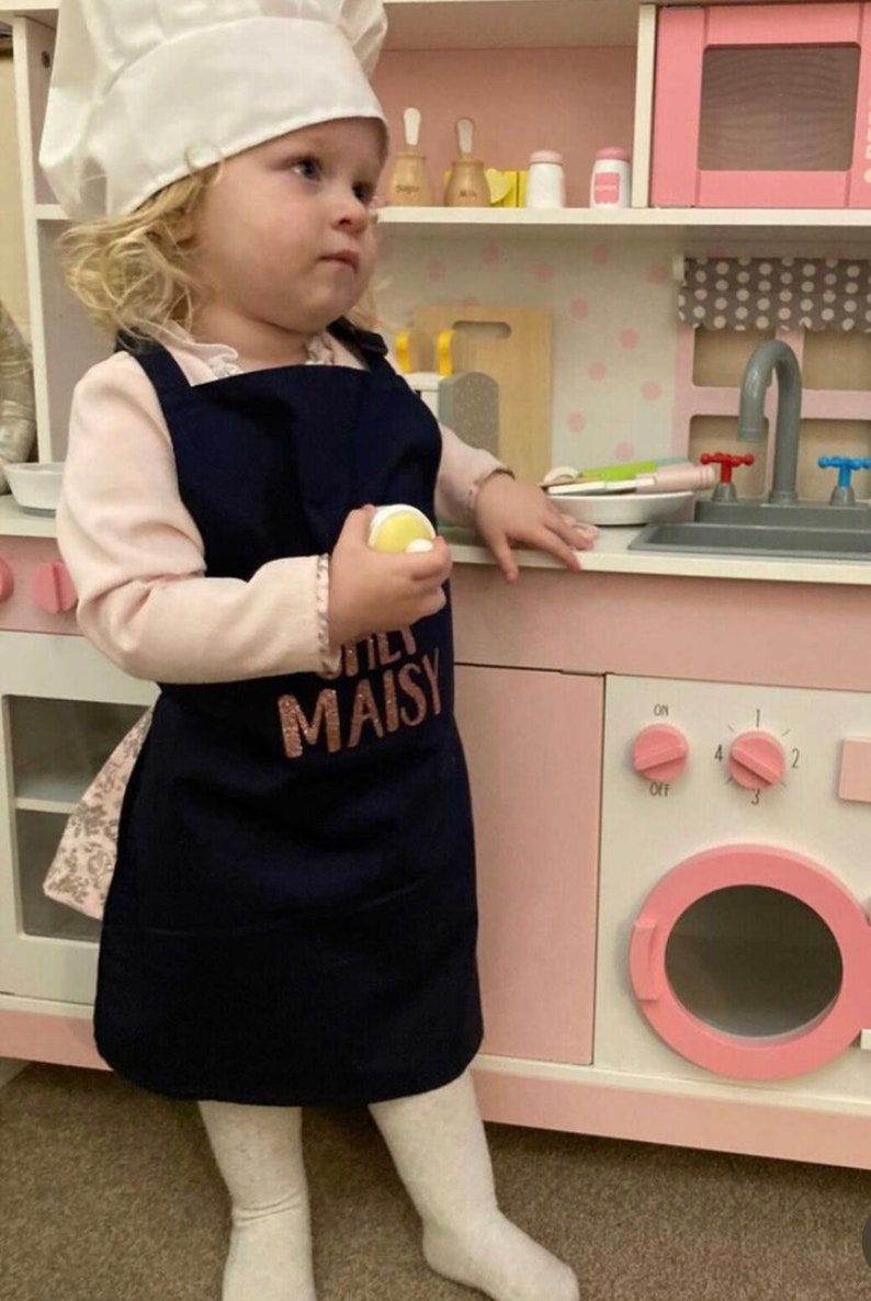 K&ouml;nnte beinhalten: Ein kleines Kind tr&auml;gt eine wei&szlig;e Kochm&uuml;tze und eine marineblaue Sch&uuml;rze mit dem Text "Little Maisy" in rosafarbenem Glitzer. Das Kind steht vor einer rosa und wei&szlig;en Spielzeugk&uuml;che.