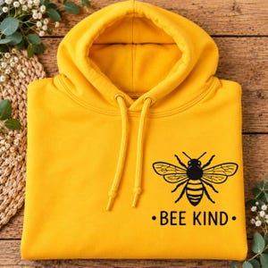 May include: A bright yellow hooded sweatshirt featuring a black graphic of a honeybee centred on the chest. Below the bee, the text "BEE KIND" is printed in a bold, black sans-serif font. The hoodie includes matching yellow drawstrings.