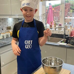 K&ouml;nnte beinhalten: Ein junger Koch tr&auml;gt eine wei&szlig;e Kochm&uuml;tze mit dem gestickten Text "CHEF GEO" und eine blaue Sch&uuml;rze mit demselben Text. Der Koch h&auml;lt in der einen Hand einen Schneebesen und in der anderen eine Edelstahl-Sch&uuml;ssel. Der Koch steht in einer K&uuml;che.