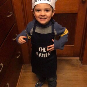 K&ouml;nnte beinhalten: Ein kleines Kind tr&auml;gt eine wei&szlig;e Kochm&uuml;tze mit dem Text "Chef Raylen" und eine blaue Sch&uuml;rze mit dem gleichen Text. Das Kind l&auml;chelt und schaut in die Kamera.