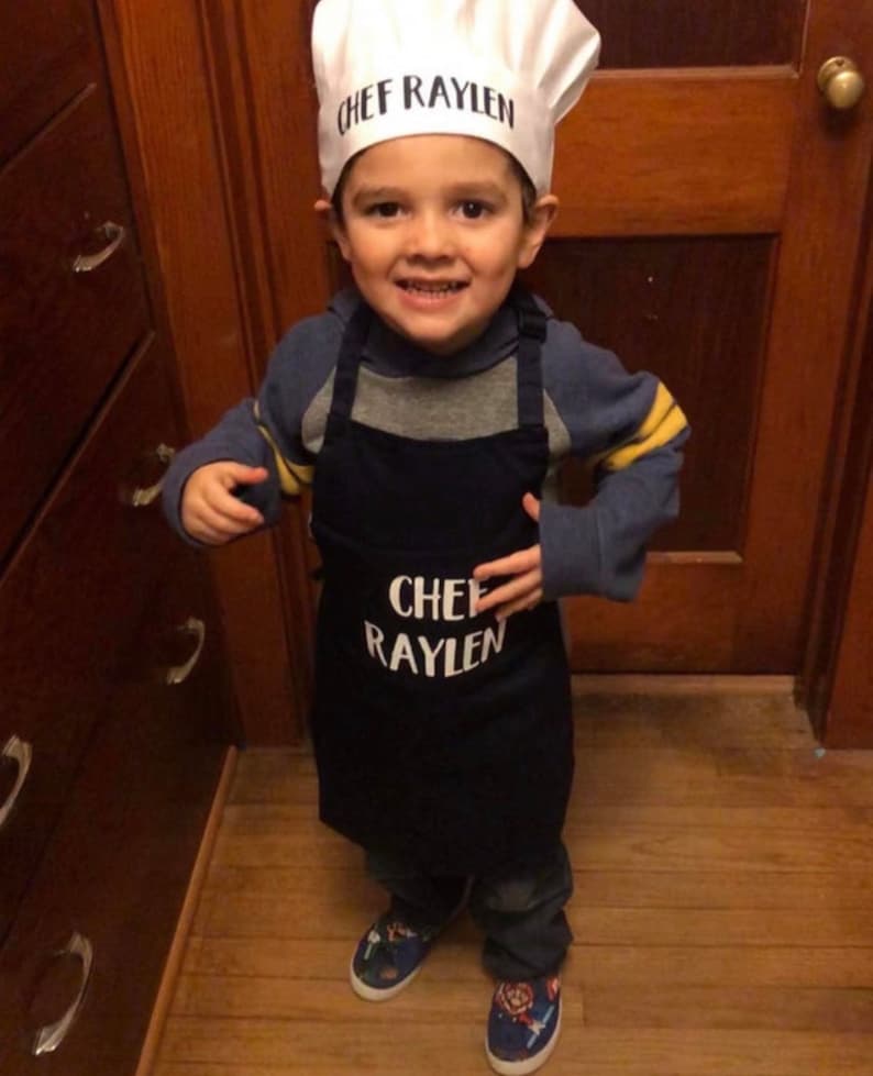K&ouml;nnte beinhalten: Ein kleines Kind tr&auml;gt eine wei&szlig;e Kochm&uuml;tze mit dem Text "Chef Raylen" und eine blaue Sch&uuml;rze mit dem gleichen Text. Das Kind l&auml;chelt und schaut in die Kamera.