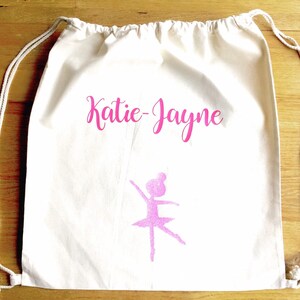 Può includere: Una borsa a cordoncino bianca con il nome "Katie-Jayne" stampato in rosa. Sulla borsa è stampata anche una silhouette rosa di una ballerina.