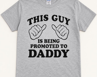 May include: A heather gray short-sleeve t-shirt featuring black text that reads THIS GUY IS BEING PROMOTED TO DADDY, accompanied by a graphic illustration of two hands with thumbs pointing toward the chest area of the shirt.
