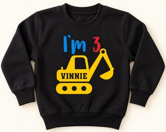 May include: A black long-sleeve sweatshirt featuring a graphic of a yellow excavator. The text "I'm" is in blue, the number "3" is in red, and the name "VINNIE" is printed in black on the side of the yellow construction vehicle.