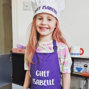 K&ouml;nnte beinhalten: Ein junges M&auml;dchen tr&auml;gt eine wei&szlig;e Kochm&uuml;tze mit dem Text "Chef Isabelle" in lila Schrift. Sie tr&auml;gt auch eine lila Sch&uuml;rze mit dem Text "Chef Isabelle" in wei&szlig;er Schrift. Die Sch&uuml;rze hat einen Wassermelonen-Druck.