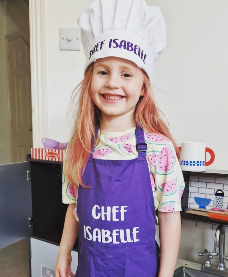 K&ouml;nnte beinhalten: Ein junges M&auml;dchen tr&auml;gt eine wei&szlig;e Kochm&uuml;tze mit dem Text "Chef Isabelle" in lila Schrift. Sie tr&auml;gt auch eine lila Sch&uuml;rze mit dem Text "Chef Isabelle" in wei&szlig;er Schrift. Die Sch&uuml;rze hat einen Wassermelonen-Druck.