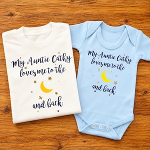 May include: A white t-shirt and a light blue baby bodysuit, both featuring the dark blue text "My Auntie Cathy loves me to the and back" with a yellow crescent moon and small gold stars printed on the front of each garment.
