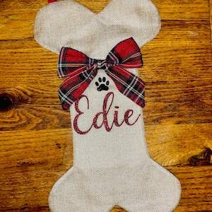 Könnte beinhalten: Ein beigefarbener Jutestrumpf in Form eines Hundebeins mit einer rot-schwarzen Karobinde und einem Pfotenabdruck. Der Strumpf ist mit dem Namen "Edie" personalisiert.