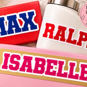 Può includere: Tre adesivi in vinile personalizzati con carattere universitario. Un adesivo blu riporta MAX su una scatola rossa, uno rosso riporta RALPH su una bottiglia bianca e uno rosa riporta ISABELLE su una scatola rosa. Un'etichetta circolare rosa reca la scritta DOUBLE LAYERED VINYLS.