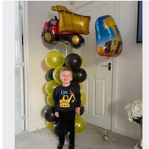 Può includere: Un bambino piccolo con una maglietta nera con un camioncino da costruzione giallo e il testo "I'm 3 Vinnie" si trova di fronte a un muro decorato con palloncini gialli e neri. Ci sono due grandi palloncini di veicoli da costruzione, uno giallo e uno blu, appesi al soffitto.