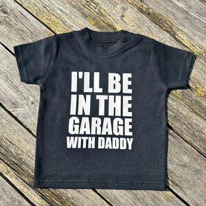 May include: Black short-sleeved t-shirt with white text that reads "I'll be in the garage with Daddy".