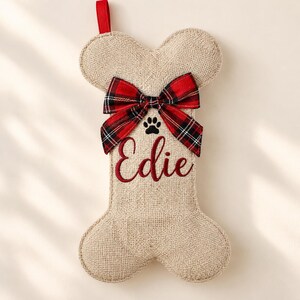 May include: A bone-shaped stocking made of tan burlap fabric featuring a red and black plaid bow at the top. The stocking is embroidered with a black paw print and the name Edie in red script. A red fabric loop is attached to the top.