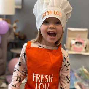 K&ouml;nnte beinhalten: Ein Kind tr&auml;gt eine wei&szlig;e Kochm&uuml;tze mit dem Text "FRESH LOU" in orangefarbenen Buchstaben. Das Kind tr&auml;gt auch eine orangefarbene Sch&uuml;rze mit dem gleichen Text in wei&szlig;en Buchstaben.