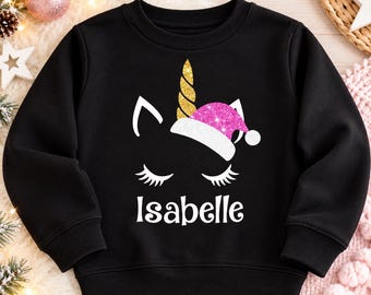May include: A black long-sleeve sweatshirt featuring a graphic of a unicorn face with closed eyes and long eyelashes. The unicorn has a gold glitter horn and wears a pink glitter Santa hat. The name Isabelle is printed in white below the graphic.