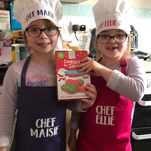 K&ouml;nnte beinhalten: Zwei junge M&auml;dchen tragen wei&szlig;e Kochm&uuml;tzen mit ihren Namen "Chef Maisie" und "Chef Ellie" und tragen lila und rote Sch&uuml;rzen. Sie halten eine Schachtel mit Keksmischung mit einer Illustration von Weihnachtsmann und dem Text "Cookie's for Santa".