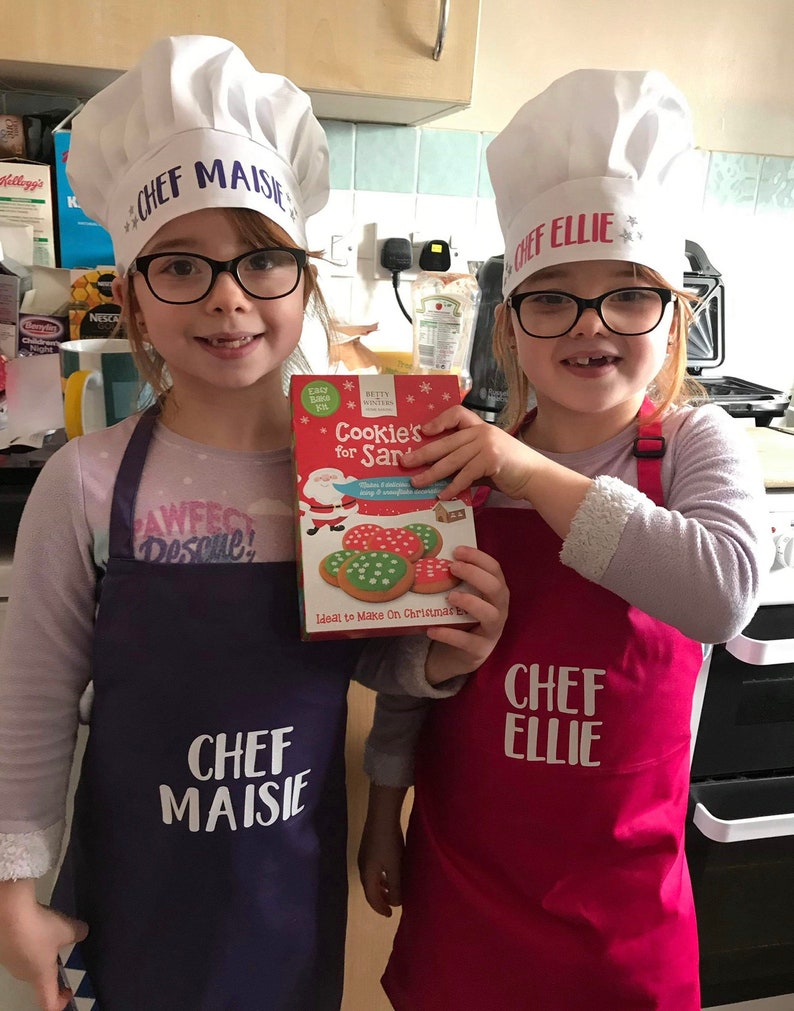 K&ouml;nnte beinhalten: Zwei junge M&auml;dchen tragen wei&szlig;e Kochm&uuml;tzen mit ihren Namen "Chef Maisie" und "Chef Ellie" und tragen lila und rote Sch&uuml;rzen. Sie halten eine Schachtel mit Keksmischung mit einer Illustration von Weihnachtsmann und dem Text "Cookie's for Santa".