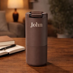 May include: A matte brown cylindrical insulated tumbler featuring the name John printed in a light-colored serif font on the upper section. The base of the tumbler has the word BRUMATE embossed vertically into the surface, and it includes a dark brown lid.