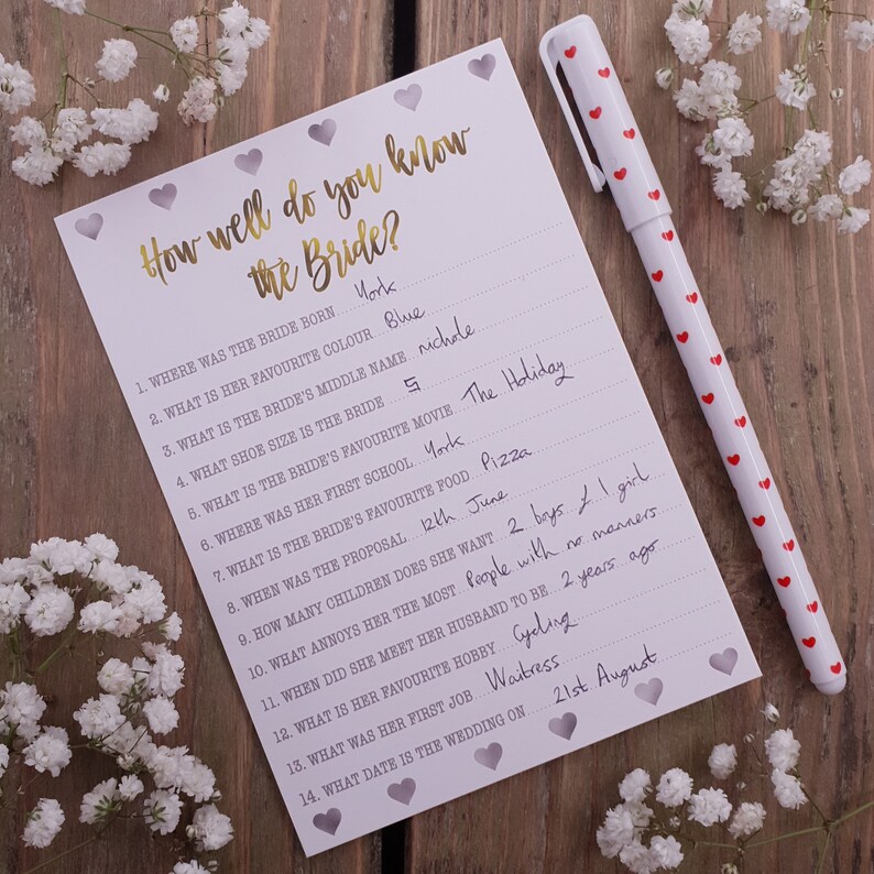 May include: A white card with a gold title "How well do you know the Bride?" and a list of questions about the bride. The card has a pink and white heart pattern and is on a wooden surface.