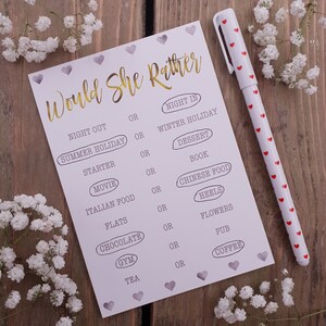 May include: A white card game with the text "Would She Rather" in gold lettering. The card features a series of questions with two choices for each question, such as "Night Out" or "Night In".