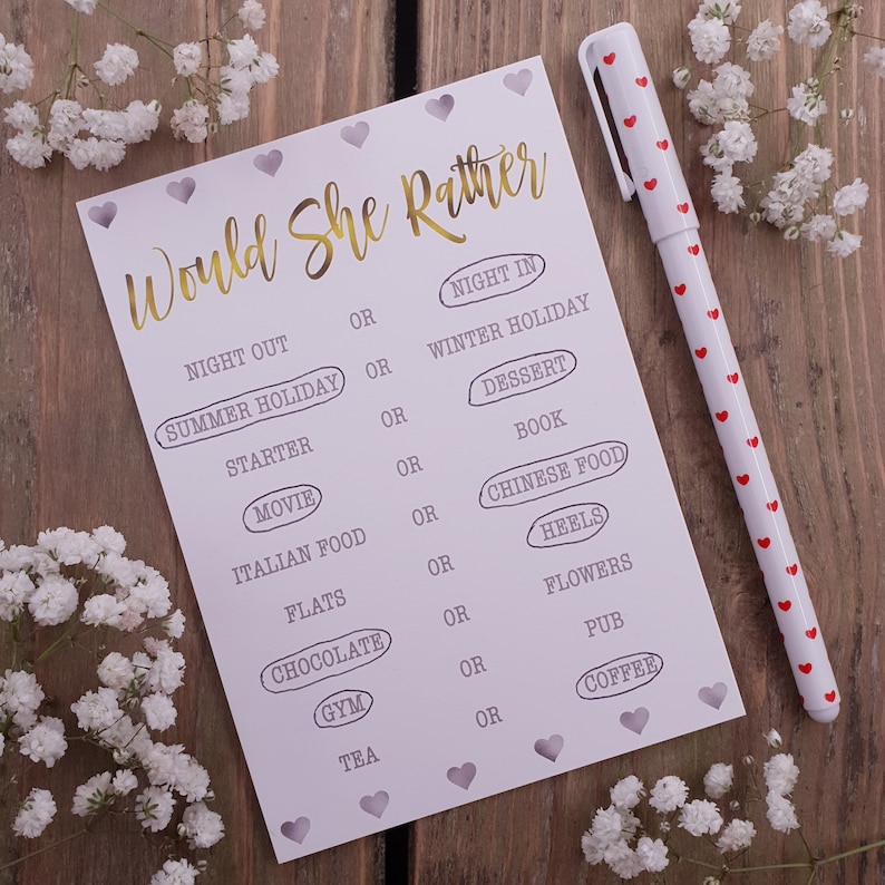 May include: A white card game with the text "Would She Rather" in gold lettering. The card features a series of questions with two choices for each question, such as "Night Out" or "Night In".