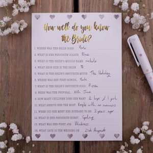May include: A white card with a gold title "How well do you know the Bride?" and a list of 14 questions about the bride with handwritten answers. The card is decorated with small purple hearts.
