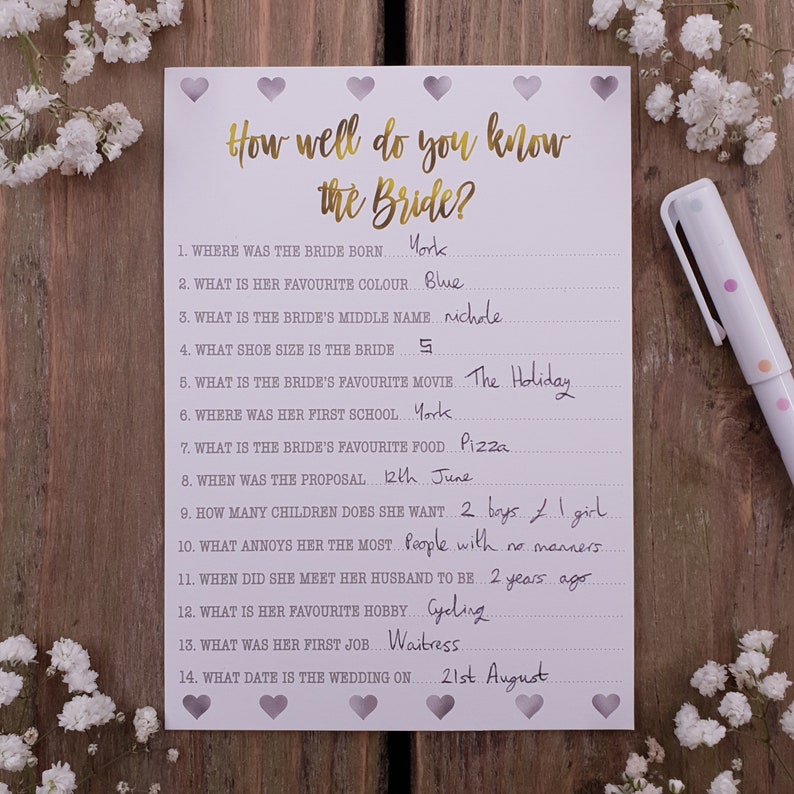May include: A white card with a gold title "How well do you know the Bride?" and a list of 14 questions about the bride with handwritten answers. The card is decorated with small purple hearts.