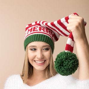 May include: A woman wearing a white fluffy sweater and a red and white striped knitted hat with a green pom-pom. The hat has the word "Merry" written on it.