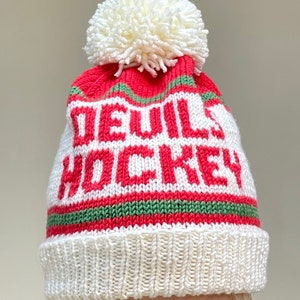 Pode incluir: Boné de malha branca com uma faixa listrada vermelha e verde e as palavras "Devils Hockey" em malha vermelha. O boné tem um pompom branco no topo.