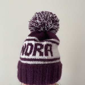 May include: Purple knitted beanie with a white pom pom and the name "ANDREA" knitted in white on the front.
