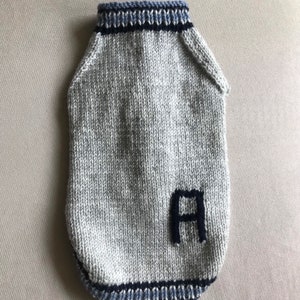 May include: A gray knitted dog sweater with blue and black trim. The sweater has a large black letter "A" knitted into the front.