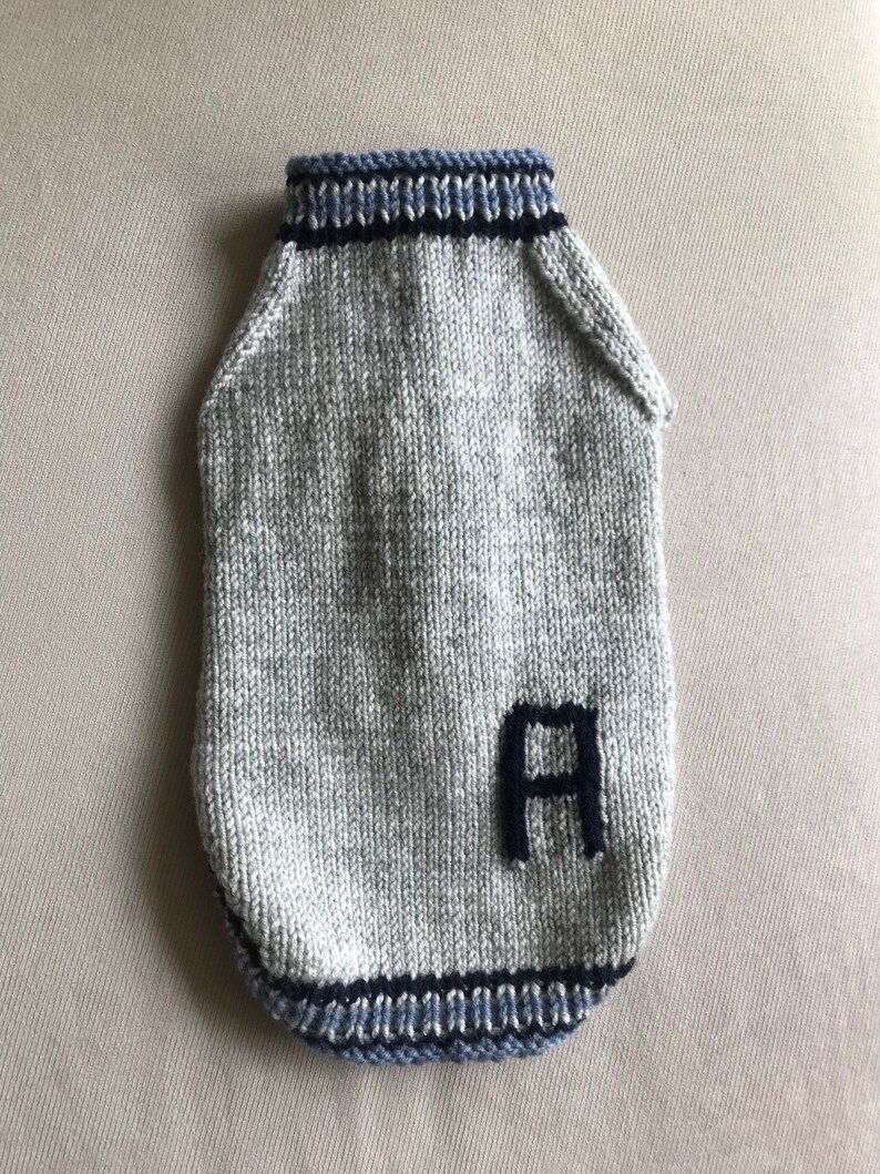 May include: A gray knitted dog sweater with blue and black trim. The sweater has a large black letter "A" knitted into the front.