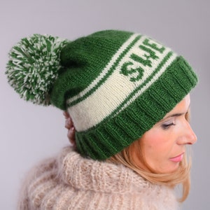 May include: A green and white knitted beanie with a pom pom on top. The beanie has the word "SWE" knitted into the white band.