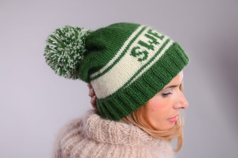May include: A green and white knitted beanie with a pom pom on top. The beanie has the word "SWE" knitted into the white band.
