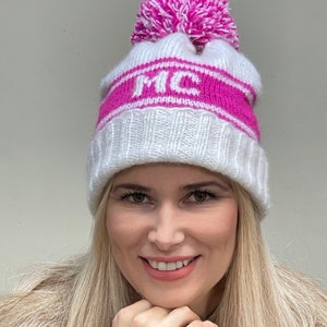 May include: White knitted beanie with a pink pom pom and a pink stripe with the letters "MC" in white.
