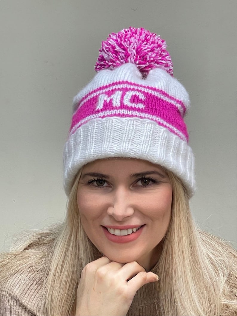 May include: White knitted beanie with a pink pom pom and a pink stripe with the letters "MC" in white.