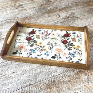 Könnte beinhalten: Ein rechteckiges Serviertablett aus Holz mit floralem Design. Das Tablett hat einen hellbraunen Holzrahmen mit ausgeschnittenen Griffen. Der Innenraum zeigt einen weißen Hintergrund mit einem farbenfrohen Blumenmuster, darunter rote Rosen, gelbe Blumen und Schmetterlinge.