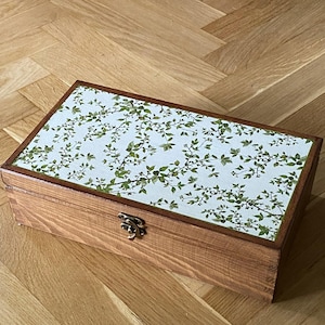 Può includere: Una scatola rettangolare in legno con coperchio incernierato caratterizzato da una superficie bianca decorata con un motivo di foglie e rami verdi. La scatola è realizzata in legno di tonalità media con venature visibili e una piccola chiusura metallica argentata.