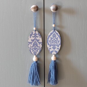 Op de afbeelding: Twee blauwe en witte ovale ornamenten met blauwe kwastjes die aan zilveren kastknoppen hangen.