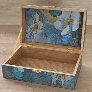 Peut inclure: Une boîte rectangulaire en bois avec un couvercle ouvert, présentant un motif floral bleu et blanc sur l'extérieur et l'intérieur du couvercle. La boîte possède des charnières, un loquet et un bras de support en métal doré, avec une base doublée de tissu beige texturé.