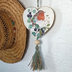 Może przedstawiać: Drewniana ozdoba w kształcie serca z rudzikiem i jagodami na białym tle. Ozdoba ma zielony frędzlem i drewniany koralik.