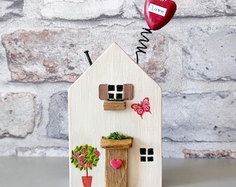 May include: A small wooden house ornament painted cream with a brown door, a window with shutters, and a painted rose bush. A black metal spring extends from the roof, topped with a red heart featuring the word love. A red butterfly is painted on the side.