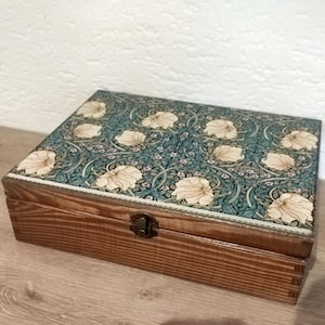 Könnte beinhalten: Eine rechteckige Holzschatulle mit Klappdeckel, der mit einem blütenartigen Muster in Blaugrün und Creme verziert ist. Die Seiten der Schatulle zeigen eine natürliche Holzmaserung, und an der Vorderseite befindet sich ein kleiner Metallverschluss.