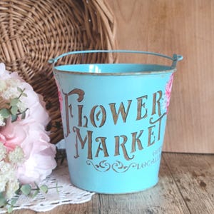 Puede incluir: Un cubo de metal azul claro con asa, con las palabras "Flower Market" en letras marrones. El cubo tiene un acabado envejecido y detalles florales. Una cesta de mimbre y flores están en el fondo.