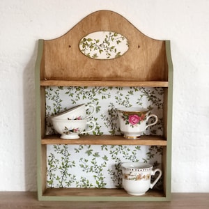 May include: A wooden wall shelf with two tiers, featuring a light green frame and a white backing with a green leaf pattern. The top shelf holds two white teacups, and the bottom shelf holds one white teacup with a floral design.