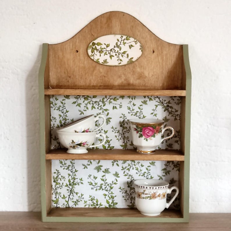 May include: A wooden wall shelf with two tiers, featuring a light green frame and a white backing with a green leaf pattern. The top shelf holds two white teacups, and the bottom shelf holds one white teacup with a floral design.
