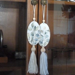 Może przedstawiać: Dwa owalne drewniane ozdoby z białym tłem i niebiesko-zielonym wzorem kwiatowym. Każda ozdoba ma biały frędzl wiszący na drewnianej koraliku.
