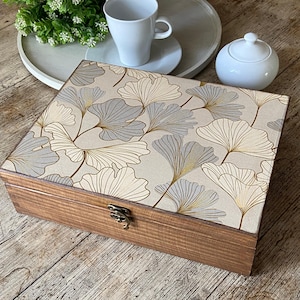 Può includere: Una scatola rettangolare in legno con coperchio incernierato caratterizzata da una superficie beige decorata con un motivo ripetitivo di foglie di ginkgo grigie e dorate. La scatola ha una finitura in legno marrone scuro e una piccola chiusura.